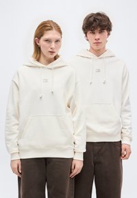 Δύο νέοι άνθρωποι στέκονται δίπλα-δίπλα φορώντας μπεζ  μπλουζάκια με κουκούλες και σκούρα καφέ παντελόνια σε απλό λευκό φόντο.