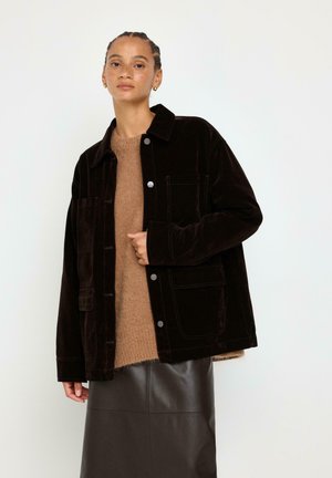 Kvinde med flettet hår iført sort knapjakke over brun sweater og mørkt lædernederdel mod ensartet lys baggrund.