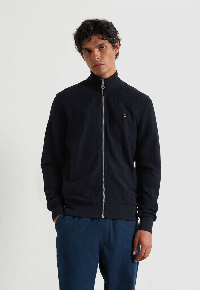 Chaqueta navy con cremallera y cuello alto, de textura acanalada, y un pequeño logotipo dorado como acento. Combinada con pantalones azules, el modelo se encuentra de pie frente a un fondo neutro.