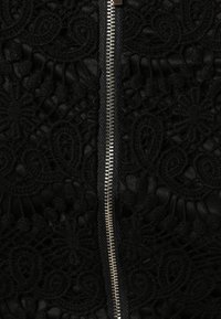 Tissu noir avec un motif en dentelle complexe, comportant une fermeture éclair métallique centrale. La texture est superposée et présente des motifs délicats.