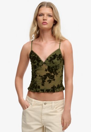 Ung kvinde med langt blondt hår iført en olivengrøn blomstret blondecamisole og cremede bukser med høj talje, stående med den ene hånd i lommen.