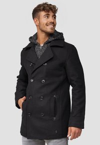 Svart dobbelknappet peacoat med store knapper, sidelommer og en grå hettegenser under. Teksturert stoff, tilpasset passform og rene linjer.
