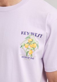 Levandulové bavlněné tričko s grafikou pomerančů a zelených listů, s texty "KEY-WEST" a "PETROL IND" v purpurových a zelených akcentových barvách.