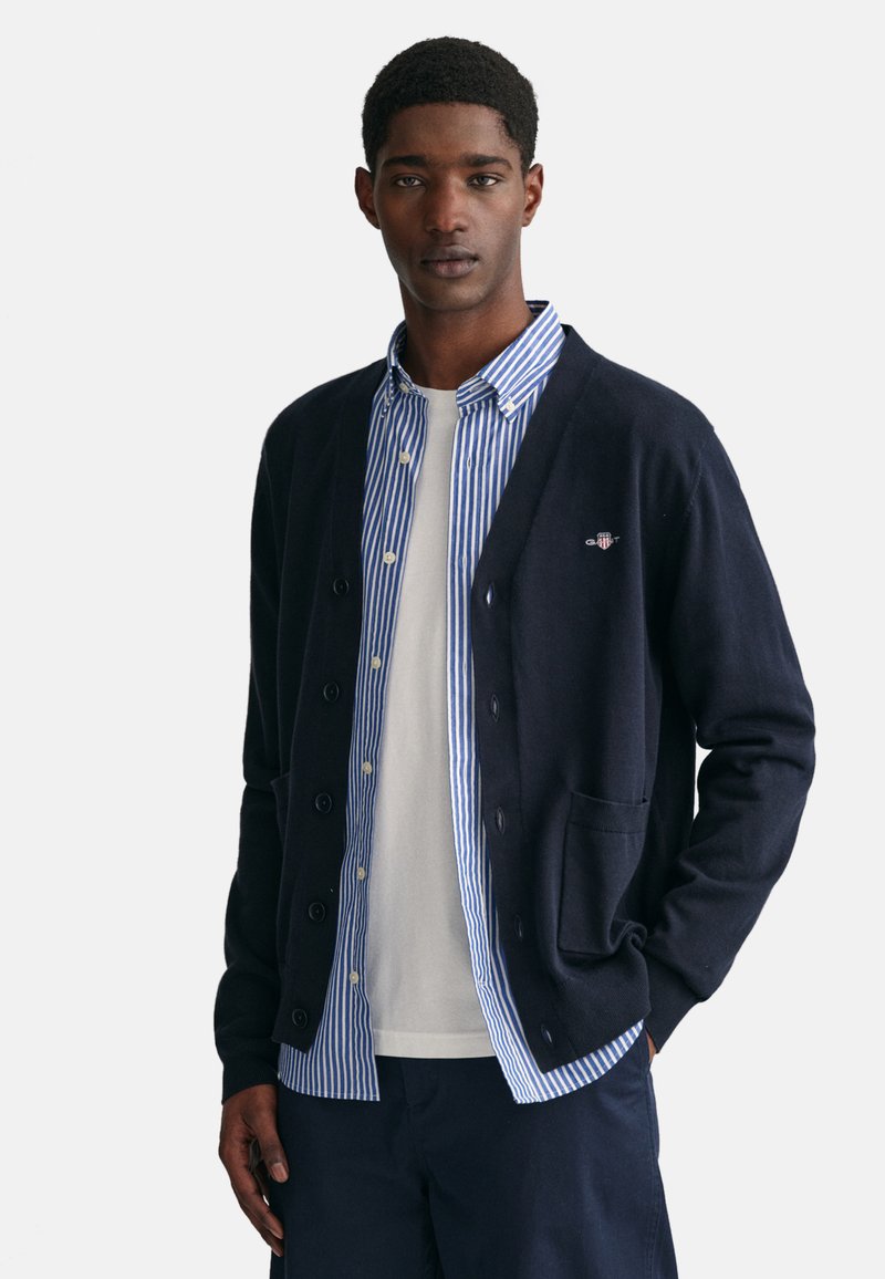 Navy-Cardigan aus weichem Stoff mit V-Ausschnitt, Knopfverschluss und Fronttaschen, über einem blau-weißen gestreiften Hemd getragen.
