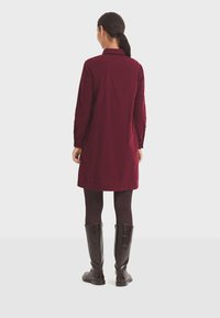 Vestido de pana burdeos con mangas largas, cuello y bolsillos laterales, combinado con botas de cuero marrón hasta la rodilla.