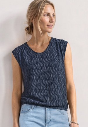 Femme aux cheveux blonds attachés porte un haut sans manches bleu marine texturé et un jean bleu clair, regardant sur le côté avec un léger sourire.