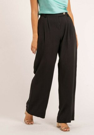 Femme portant un pantalon noir taille haute à jambes larges et des sandales à talons blocs dorées à bout ouvert, debout devant un fond clair uni.