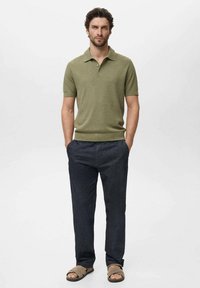 Polo shirt vert olive à manches courtes avec un design tricoté texturé, associé à un pantalon foncé et des sandales beiges, présenté sur un fond clair.