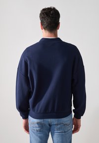 Jersey azul marino con una textura en la tela, corte relajado y puños y dobladillo acanalados. Llevado con jeans de color azul claro, mostrando la vista trasera.