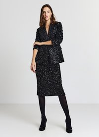 Schwarzer Blazer- und Rock-Set mit Pailletten, strukturierter Oberfläche, tiefem V-Ausschnitt und langen Ärmeln, kombiniert mit schwarzen High Heels und Strumpfhosen.