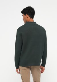 Mørkegrønn genser med lange ermer, krage og en avslappet passform. Glatt tekstur, bakfra, sammen med beige kordfløyelsbukser.