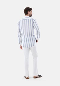 Camicia di lino con righe verticali blu e bianche, maniche lunghe, polsini con bottone e orlo arrotondato; abbinata a pantaloni bianchi e sandali casual.