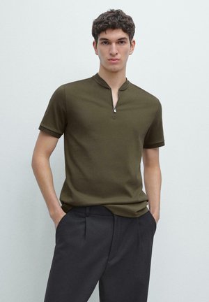 Junger Mann steht mit Händen in den Taschen, trägt ein olivgrünes, kurzärmeliges Hemd mit Reißverschluss am Kragen und schwarze Hose vor einem neutralen Hintergrund.