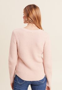 Helles rosa, geripptes Sweatshirt mit langen Ärmeln, V-Ausschnitt und abgerundetem Saum, kombiniert mit blauen Jeans. Die Textur ist weich und gestrickt.
