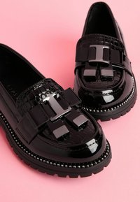 Loafers din patent negru, cu un model texturat de crocodil și o fundă decorativă mare. Talpa groasă este accentuată cu ținte argintii. Finisaj neted.
