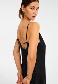 Schwarzes Satin-Rutschkleid mit dünnen Schulterträgern, offenem Rücken und seitlichen Schlitzen. Das Model trägt funkelnde Ohrringe und hat einen neutralen Hintergrund.