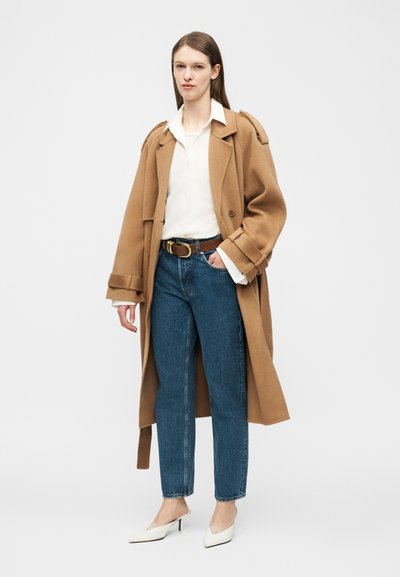 Donna che indossa un cappotto color cammello sopra una camicia bianca, jeans blu, cintura marrone e tacchi bianchi con punta affilata, in piedi davanti a uno sfondo semplice.