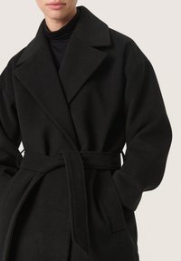 Cappotto di lana nera con vita regolabile, ampie rever e spalle pronunciate. Presenta maniche lunghe e tasche laterali, texture liscia.
