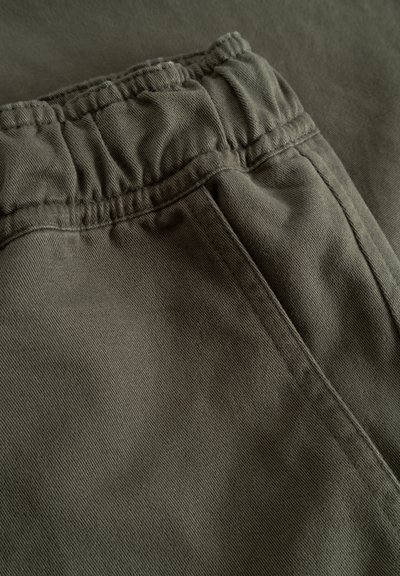 Gros plan sur un tissu vert olive avec une ceinture élastique et des coutures, probablement une partie d'un pantalon ou d'un short décontracté.