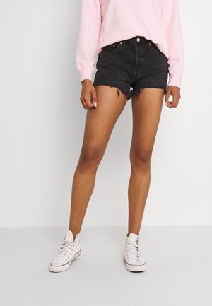 Shorts en denim noir taille haute avec ourlet effiloché, associés à une chemise à manches longues rose clair et des baskets montantes blanches.