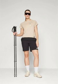 Light beige t-shirt with a small pocket, black shorts, and white sneakers. Accessories include a hiking pole and dark sunglasses.