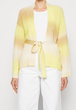 Strikket cardigan i gradient fra gul til crème, med et bælte, åben front og ribbede manchetter. Båret over en hvid top og lyse bukser.