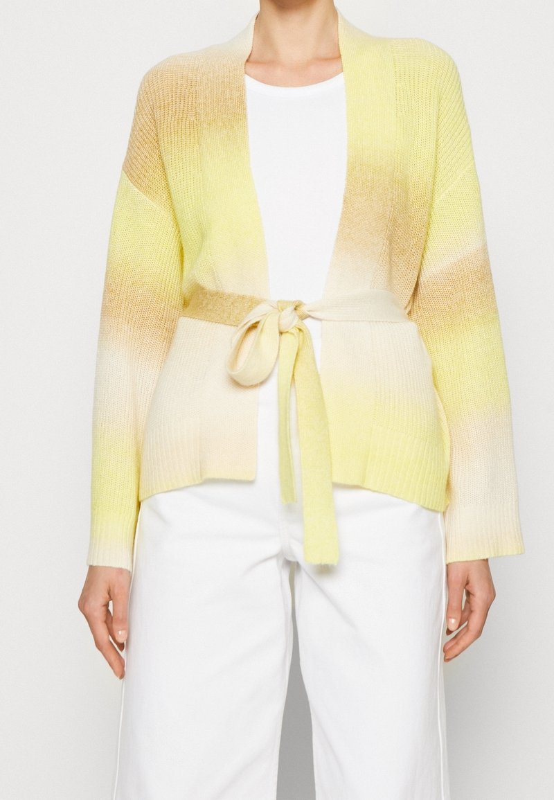 Cárdigan de punto en degradado amarillo a crema, con cinturón atado, frente abierto y puños acanalados. Llevado sobre una blusa blanca y pantalones claros.