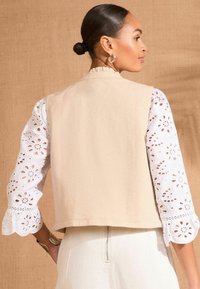 Beige cropped vest with a scalloped neckline, paired with white broderie anglaise sleeves featuring floral cutouts and ruffled cuffs.