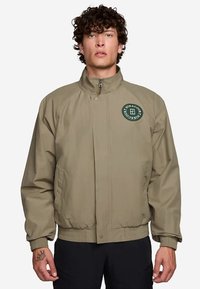 Khaki bomber jacket with a high collar, ribbed cuffs, and a large round logo on the chest. Fabric appears smooth and lightweight.
