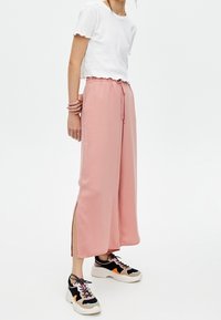 Femme portant un pantalon large rose clair avec des fentes sur les côtés, un haut court à manches courtes blanc, et des baskets multicolores à lacets.