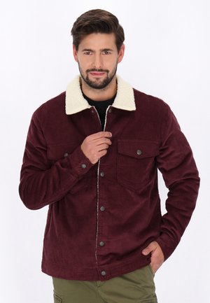 Bărbat purtând o jachetă din catifea côtelé burgund cu guler alb de fleece, ținând partea din față a jachetei cu o mână, stând în fața unui fundal alb simplu.