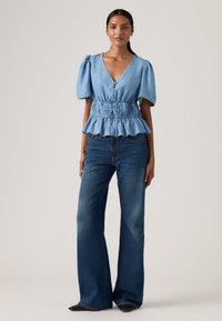 Ανοιχτό μπλε τοπ με φουσκωτά μανίκια, βαθύ V ντεκολτέ, σούρα στη μέση και βολάν στο τελείωμα, συνδυασμένο με σκουρό μπλε τζιν φαρδιά στο κάτω μέρος και παπούτσια με μυτερή μύτη.