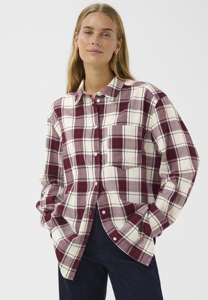 Giovane donna con lunghi capelli biondi, che indossa una camicia a quadri rosso e bianco con bottoni e pantaloni scuri, che sta in piedi con le mani nelle tasche.