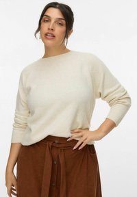 Cremefarvet, blødt strikket sweater med raglanærmer. Kombineret med en brun fløjlskørt med bundet talje og knapdetaljer.