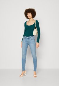 Teal langermet bodysuit med rynket halslinning, kombinert med lyseblå slim-fit jeans. Tilbehør inkluderer en liten hvit håndveske og høyhælte sandaler.