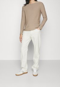 Chemise à manches longues en coton marron clair, associée à un pantalon blanc et des baskets blanches avec des accents gris, créant un look décontracté monochrome.