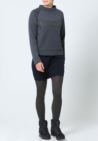 Donna che indossa un felpa grigio scuro con la scritta "FIRE", una gonna blu navy, collant grigi e scarponi da trekking neri, in piedi contro uno sfondo semplice.