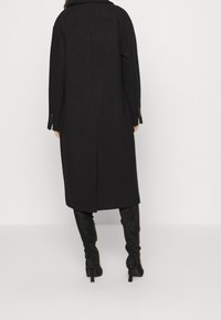 Manteau long noir en mélange de laine, présentant une silhouette droite, des poches latérales et des poignets boutonnés. Texture lisse avec un design minimal.