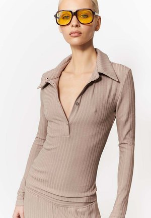 Kobieta w beżowym prążkowanym topie z długim rękawem i głębokim dekoltem w kształcie litery V oraz dużych kwadratowych okularach przeciwsłonecznych z żółtymi szkłami na białym tle.