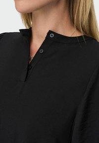 Czarna koszula z kołnierzykiem, z zapięciem na guziki, o miękkiej teksturze i lekko bufiastej krótkiej rękaw. Prosty, minimalistyczny design.