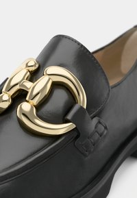 Zwarte leren loafers met een prominente glanzende gouden metalen paardenbitdetail op de voet, soepele stiksels en een lage gestapelde hak.