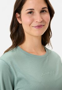 Hellgrünes T-Shirt mit kurzen Ärmeln aus weichem Stoff, mit Rundhalsausschnitt und prägtem Logo auf der Brust. Geschmeidige Textur, lässiges Design.