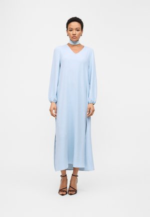 Frau, die ein hellblaues, langärmliges, knöchellanges Kleid mit passendem Choker trägt, steht mit den Armen seitlich am Körper und trägt schwarze Absätze.