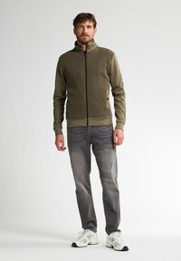 Olijfgroene ritsjack met een gestructureerde bovenkant, lange mouwen en geribde manchetten. Gecombineerd met grijze denim jeans en witte sneakers.