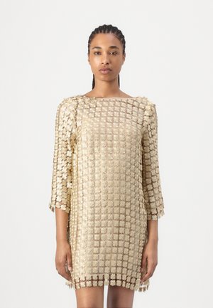 Femme aux cheveux tressés porte une robe dorée semi-transparente avec des motifs floraux texturés et des manches trois-quarts, debout sur un fond blanc.