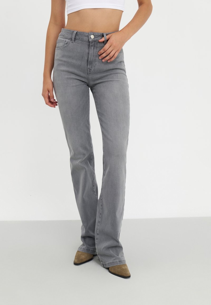 Jean gris à taille haute et coupe ajustée, porté avec des chaussures marron à bout pointu et un top blanc court, mains détendues près de la taille.