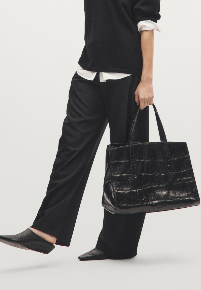 Borsa tote nera con stampa in cocco, manici piatti, forma rettangolare e superficie liscia, abbinata a pantaloni neri su misura e scarpe slip-on nere.