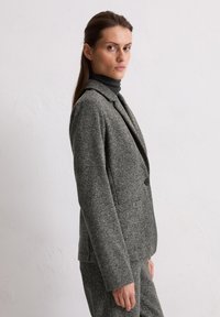 Grijze, gestructureerde blazer met een revers, een enkele knoopsluiting en twee voorzakken, gecombineerd met een donkere coltrui.