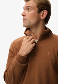Brauner Reißverschluss-Pullover mit geripptem Textur und hohem Kragen. Verfügt über ein kleines Logo auf der Brust und eng anliegende Bündchen. Glatte Stoffoberfläche.