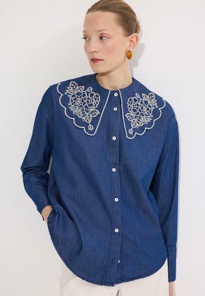 Žena oblečená v modrej košeli s gombíkmi a veľkým bielym vyšívaným kvetinovým golierom, ruky v vreckách, pozerá sa do strany na bielom pozadí.